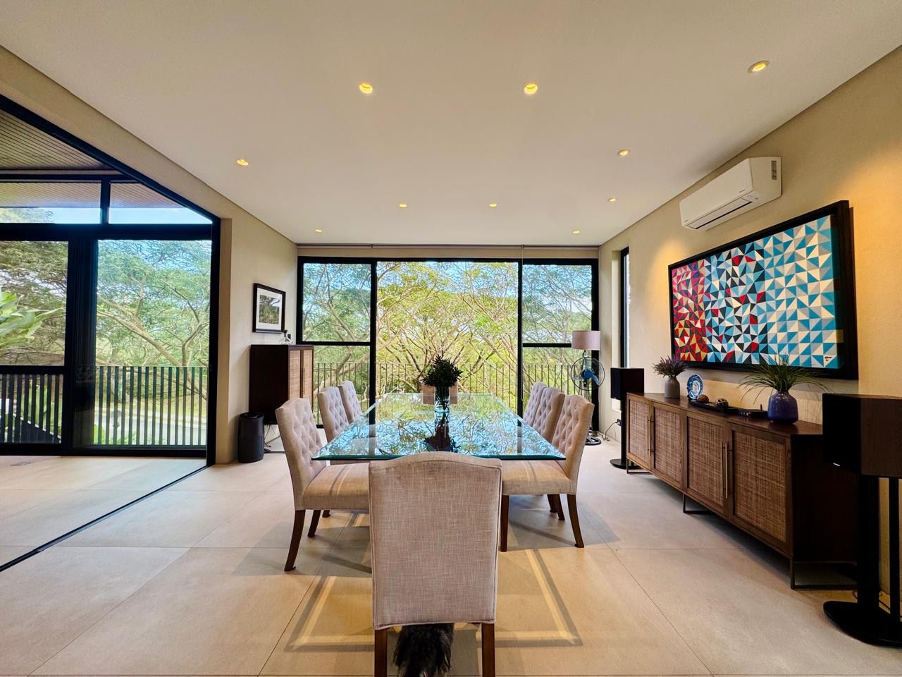 Formal dining room with glass-top table and tufted chairs, opening to forest views through floor-to-ceiling glass